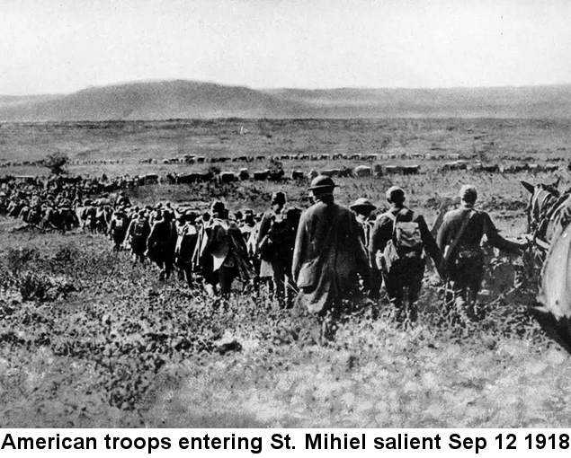 Black and white photo of hundreds of soldiers on foot and on horseback, along with trucks and horse-drawn wagons, marching in a single zig-zagging line across a broad plain, toward a round-topped hill in the distance. 